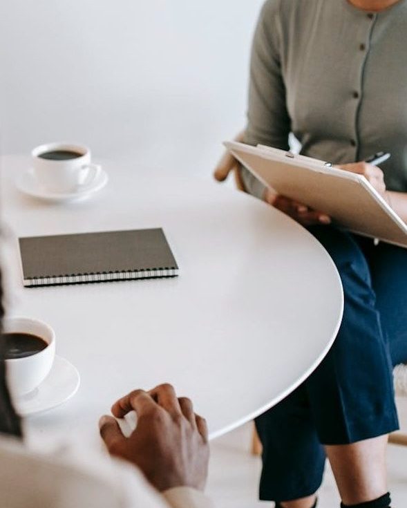 Professional ethnic female psychotherapist asking questions to black male patient and taking notes in clipboard while sitting together at table in modern psychotherapy center
